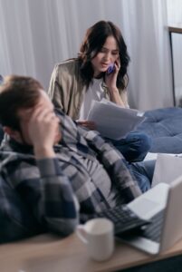 Worried couple handling bills and taxes at home, seated on a sofa, discussing on the phone.